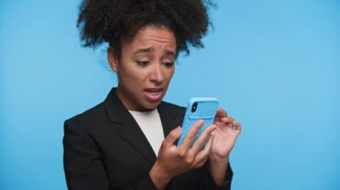 Spor ceketli genç bir kadın şok olmuş gibi görünüyor ve mavi arka planda izole edilmiş akıllı telefonundan üzücü ya da şaşırtıcı haberler okurken ağzını kapatıyor..