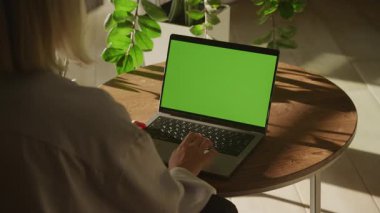 Bir kadın yeşil ekranlı bir dizüstü bilgisayarda güneşli bir odada bitkilerle birlikte tahta bir masada otururken....