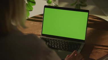 Bir kadın ahşap bir masada yeşil ekran bir dizüstü bilgisayara bakıyor, sıcak bir şekilde aydınlatılmış kapalı bir alanda bitkilerle çevrili..