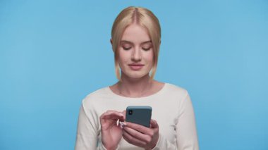 Akıllı telefonuyla kayarken gülümseyen genç bir kadın stüdyo ortamında mavi bir arka planın önünde dikiliyor..