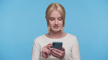 Akıllı telefonuyla uğraşan genç bir kadın stüdyo ortamında temiz, mavi bir arka plana karşı duruyor..