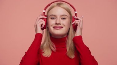 Kırmızı kulaklıklı, pembe arka planda müzik dinlemekten hoşlanan, kırmızı balıkçı yaka kazak giyen, gülümseyen genç bir kadın..