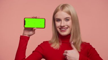 Kırmızılı, gülümseyen genç bir kadın pembe arka planda yeşil ekranlı akıllı bir telefonu tutarken baş parmağını kaldırıyor..