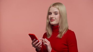 Kırmızı elbiseli, kendine güvenen, kameraya gülümseyen ve pembe arka planda akıllı telefonunu kaydıran genç bir kadın..