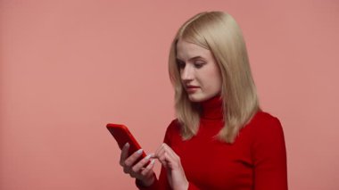 Kırmızı elbiseli genç bir kadın, pembe arka planda kötü haberler gördükten sonra geniş gözlü ve ağzı açık bir akıllı telefon tutuyor..