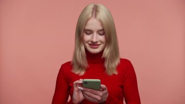 Kırmızılı mutlu genç kadın akıllı telefonuna bakıyor ve gülümsüyor, pembe arka planda online alışverişin keyfini çıkarıyor..