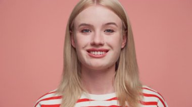 Sarı saçlı, kırmızı ve beyaz çizgili gömleğini pembe arka planda gezdirirken gülümseyen neşeli genç bir kadının yakın plan portresi..