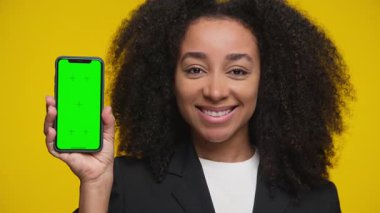 Kıvırcık saçlı mutlu kadın yeşil ekran gösteren bir akıllı telefon tutuyor, sarı arka planda uygulama sunumu için ideal.