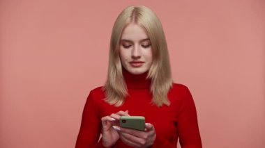 Kırmızı balıkçı yaka kazaklı sarışın bir kadın akıllı telefonundan süzülüp yumuşak pembe bir stüdyo arka planının önünde duruyor..