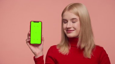 Kırmızı balıkçı yakalı mutlu sarışın kadın pembe arka planda yeşil ekranlı akıllı telefonu sunar..