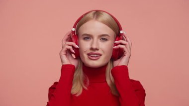 Kırmızı balıkçı yaka kazaklı sarışın kadın kulaklığını hafif kaldırır. Tuhaf bir ifadeyle, pembe arka planda dikkatle dinler..