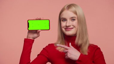 Kırmızı balıkçı yaka kazaklı sarışın kadın yeşil ekranlı akıllı bir telefona gülümsüyor ve pembe arka planda içeriği teşvik ediyor..