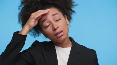 Hasta, genç, profesyonel bir kadın alnına kapalı gözlerle dokunuyor, hastalık ya da ateş belirtileri gösteriyor, mavi arka planda izole edilmiş.