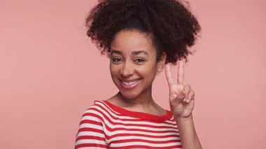 Neşeli genç bir kadın parlak bir şekilde gülümsüyor ve parmaklarıyla pembe bir arka plana barış işareti yapıyor..