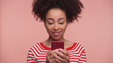 Genç bir kadın telefonuna bakıyor ve mesajlaşırken gülümsüyor. Sıradan bir kıyafetin içinde pembe bir arka plana karşı duruyor..