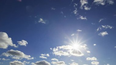 Parlak zaman dilimi, güneşin mavi bir gökyüzünde parladığını gösteriyor. Dağılmış beyaz bulutlar çerçevenin üzerinde yavaşça sürükleniyor..