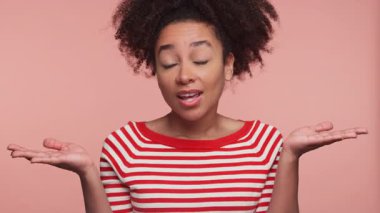 Kırmızı çizgili tişörtlü genç bir kadın omuzlarını kaldırarak omuzlarını silkiyor ve pembe bir arka plana karşı duran şaşkın bir gülümseme içinde..