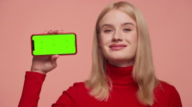 Kırmızı balıkçı yakalı, gülümseyen genç bir kadın yeşil ekranlı akıllı bir telefonu pembe arka planda tutarken onayıyla başını sallıyor..