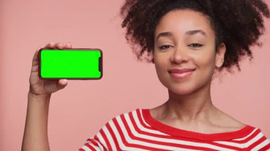 Çizgili tişörtlü neşeli genç bir kadın pembe arka planda yeşil ekranlı akıllı bir telefonu tutarken başını sallıyor..