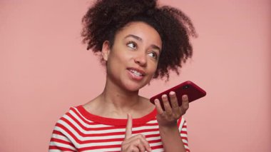 Kırmızı çizgili gömlekli genç bir kadın akıllı telefonu yatay olarak tutuyor ve ona doğru konuşuyor. Pembe arka planda bir sesli mesaj kaydediyor..