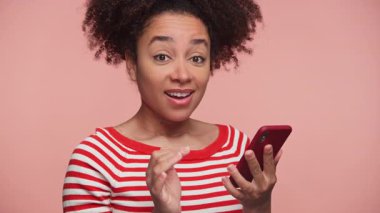 Kırmızı çizgili tişörtlü neşeli genç kadın elinde akıllı telefon tutarken şaşırmış ve heyecanlanmış görünüyor, pembe arka plana karşı olumlu haberlere tepki veriyor..