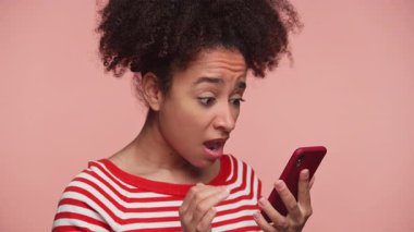 Çizgili gömlekli hayal kırıklığına uğramış genç bir kadın akıllı telefonuna şaşırmış ve hayal kırıklığına uğramış görünüyor. Pembe arka planda izole edilmiş, olumsuz bilgilere güçlü bir tepki veriyor..