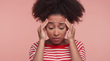 Kırmızı çizgili tişörtlü genç bir kadın. Şakaklarını kederli bir ifadeyle tutuyor. Pembe arka planda baş ağrısı çekiyor..