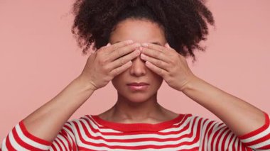 Kırmızı çizgili gömlekli genç bir kadın iki eliyle gözlerini kapatıyor. İnkâr, kaçınma ya da pembe arka plana karşı oynaşmayı ifade ediyor..