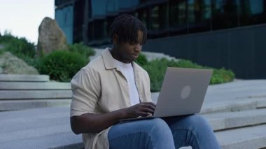 Modern basamaklarda dizüstü bilgisayarla oturan genç adam profesyonel bir şehir merkezinde ofis binasının dışında çalışıyor..