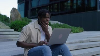 Dışarıda dizüstü bilgisayarla oturan genç adam, online alışveriş veya finansal işlem hakkında karar verirken tereddüt ve şüphe ifade ediyor..