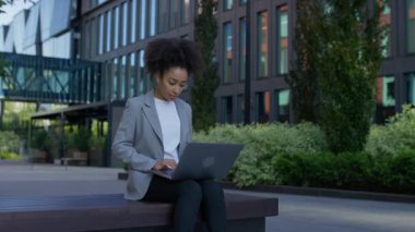 Modern şehir bölgesinde bir bankta oturan odaklanmış genç bir kadın, sıradan bir iş kıyafetiyle bilgisayarında kayıyor ya da internette geziniyor..