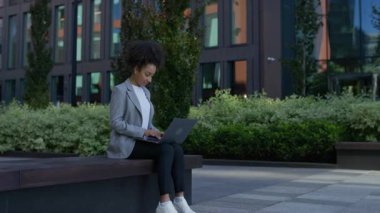 Genç profesyonel bir kadın dizüstü bilgisayarla bankta oturuyor. Etrafı ofis binalarıyla çevrili, sessiz, modern bir şehir parkında çalışmaya odaklanmış..