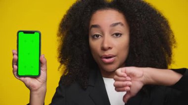 İş elbisesi giymiş, yeşil ekranlı telefonu gösteren ve baş parmağını kaldıran ciddi bir kadın, mobil uygulama veya aygıtla ilgili memnuniyetsizliğini dile getiriyor..
