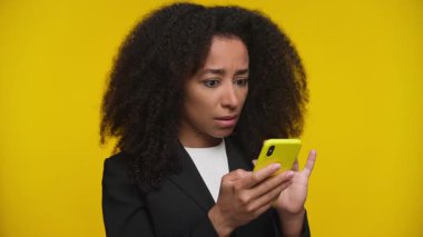 Sarı akıllı telefonundaki kötü habere duygusal tepki veren ceketli, şok olmuş ve hayal kırıklığına uğramış bir kadın cesur sarı bir arka plana karşı çıkıyor..