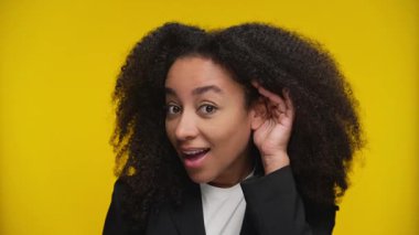 Siyah ceketli genç bir kadın, neşeli bir ifadeyle eğiliyor, sarı arka plana karşı daha dikkatli dinlemek için kulağını kaldırıyor..