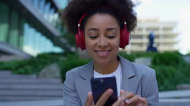 Kırmızı kulaklık takan gülümseyen kadın iş kıyafetleri içinde şehir basamaklarında otururken akıllı telefonuyla müzik dinliyor..