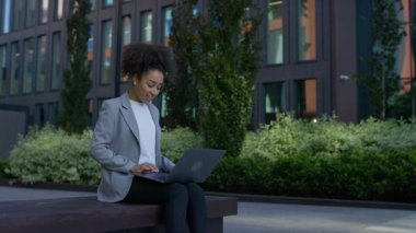 Genç bir kadın, modern bir şehir manzarasında, bir bankta otururken, dışarıda rahat bir şekilde çalışırken, dizüstü bilgisayarını kaydırıyor..