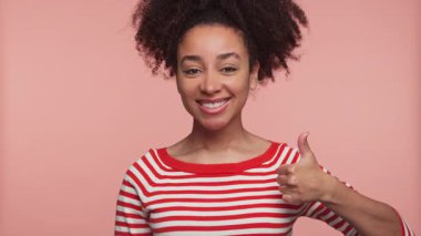 Kırmızı çizgili tişörtlü mutlu genç kadın pembe arka plana karşı pozitifliğini ifade eden neşeli bir gülümsemeyle baş parmağını kaldırıyor..