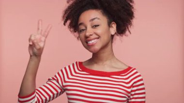 Kırmızı çizgili tişörtlü neşeli genç bir kadın yumuşak pembe bir arka plana gülümserken eliyle barış işareti yapıyor..