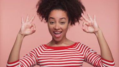 Kırmızı çizgili tişörtlü neşeli genç kadın iki el hareketi yapıyor ve pembe arka planda geniş bir gülümseme sergiliyor..