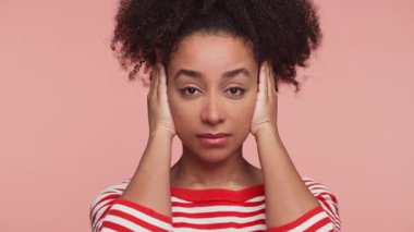 Kırmızı çizgili gömlekli genç bir kadın iki eliyle kulaklarını kapatıyor. Pembe arka planda rahatsız edici ya da reddedici bir şey gösteriyor..