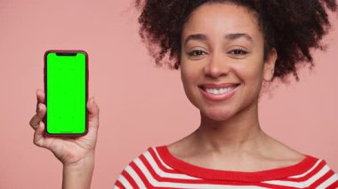 Kırmızı çizgili tişörtlü genç bir kadın yeşil ekranlı akıllı telefonu gösterirken gülümsüyor. Uygulama modeli veya UI tasarımı için mükemmel. Pembe arka planda izole edilmiş..