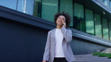 Genç bir kadın, mutlu bir telefon görüşmesi yaparken modern binanın yanında yürüyor. Gülümsüyor, rahatlamış ve nişanlı görünüyor..