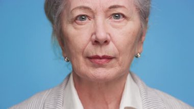 Ciddi yaşlı kadın doğrudan kameraya bakıyor, mavi arka planda resmi kıyafetler giyiyor, bilgeliği, yaşlanmayı ve kendine güveni temsil ediyor..