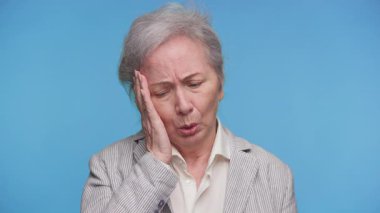 Sıkıntılı, yaşlı bir kadının yüzünü hüsran içinde tutması, artan sağlık hizmetleri veya sigorta giderleri ile ilgili endişeleri sembolize ediyor, mavi arka planda izole edilmiş..