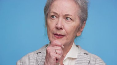 Endişeli ve düşünceli görünen yaşlı bir kadın, mavi arka planda izole edilmiş, önemli sağlık hizmetleri veya sigorta seçenekleri hakkında düşünmeyi veya belirsizliği sembolize ediyor..