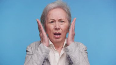 Şaşırmış ve paniğe kapılmış yaşlı bir kadın, mavi arka planda izole edilmiş, beklenmedik sağlık ve finans haberleri nedeniyle şok ya da korkuyu temsil ediyor..