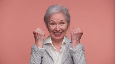 Neşeli, yaşlı bir kadın geniş bir gülümseme ve kutlama için iki elini de kaldırıyor. Başarı, coşku ve pembe arka planda izole edilmiş bir yaşam anını ifade ediyor..