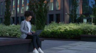 Ofis binasının dışında bankta oturan genç profesyonel bir kadın, modern bir şehir ortamında dizüstü bilgisayarla yazarken konsantre oluyor..