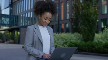 Dışarıda dizüstü bilgisayar kullanan, düşünceli bir şekilde gülümseyen ve duraklayan, üretkenliği, yaratıcılığı ve modern çalışma tarzını sembolize eden genç profesyonel kadın..